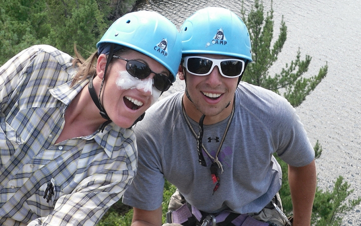 Two people wearing helmets smile at the camera. They appear to be at a high elevation. There is water below them.